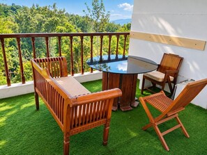 A balcony with a round table and wooden chairs, surrounded by artificial grass, overlooking a forested landscape.