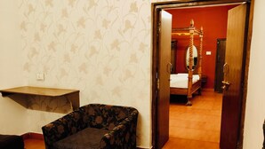 A room with a wooden floor, a corner sofa, a small table, and a door leading to a bedroom with a four-poster bed and a mirror.
