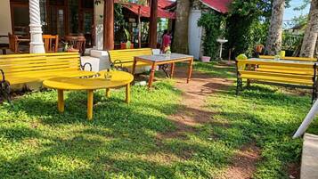 An outdoor seating area with yellow tables and benches, surrounded by greenery and a building with a tiled roof.
