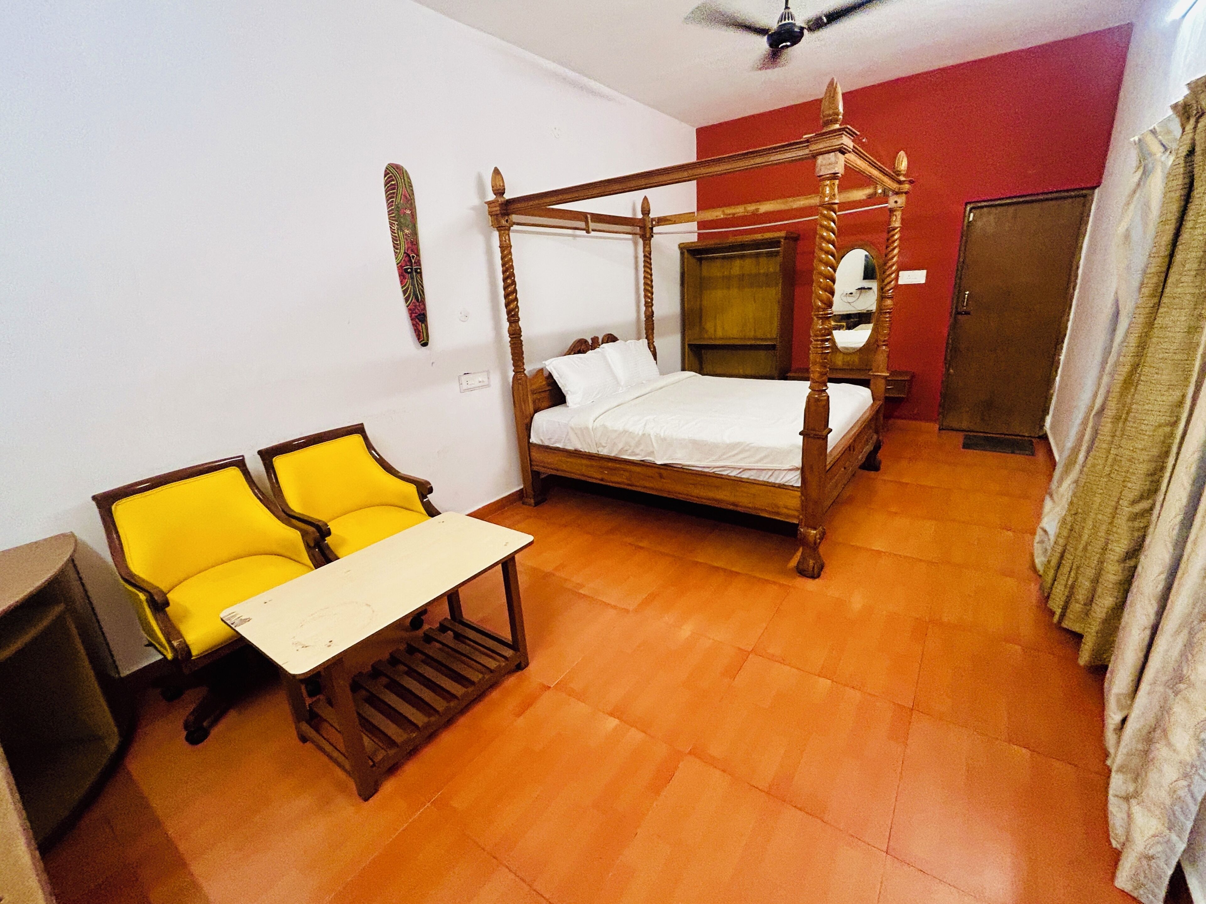 A room with a four-poster bed, a yellow armchair, a small table, and a ceiling fan.