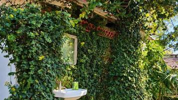 Un jardín con un lavamanos blanco sobre pedestal, un tazón rosa y un espejo en una pared cubierta de follaje verde.