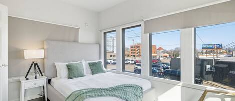 A modern bedroom with a large bed, a bedside table with a lamp, and a view of a parking lot through large windows.