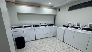 A laundry room with multiple washing machines and dryers, a trash bin, and a grey tiled floor.