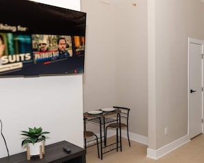 A hallway with a TV mounted on the wall displaying an advertisement, a small table with chairs, and a potted plant. - 610 Large Studio Downtown With Pool Table (Albany)