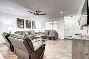 Un salon spacieux comprenant un canapé, deux fauteuils, une table basse et un ventilateur de plafond. Les murs sont ornés de tableaux encadrés et une fenêtre est équipée de stores.