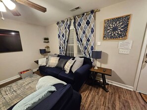 A living room with a dark blue sofa, two side tables with lamps, a patterned curtain, a wall-mounted TV, and a decorative wall piece.