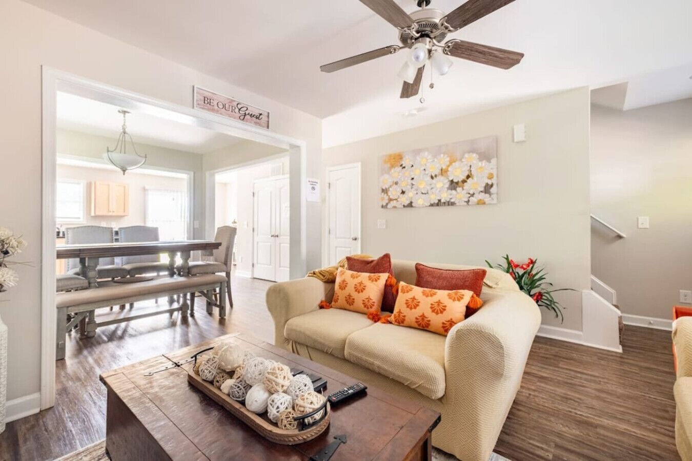 A spacious living room with a beige sofa, a coffee table with decorative items, a ceiling fan, and a dining area with a chandelier.