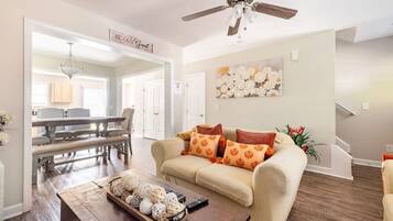 A spacious living room with a beige sofa, a coffee table with decorative items, a ceiling fan, and a dining area with a chandelier.