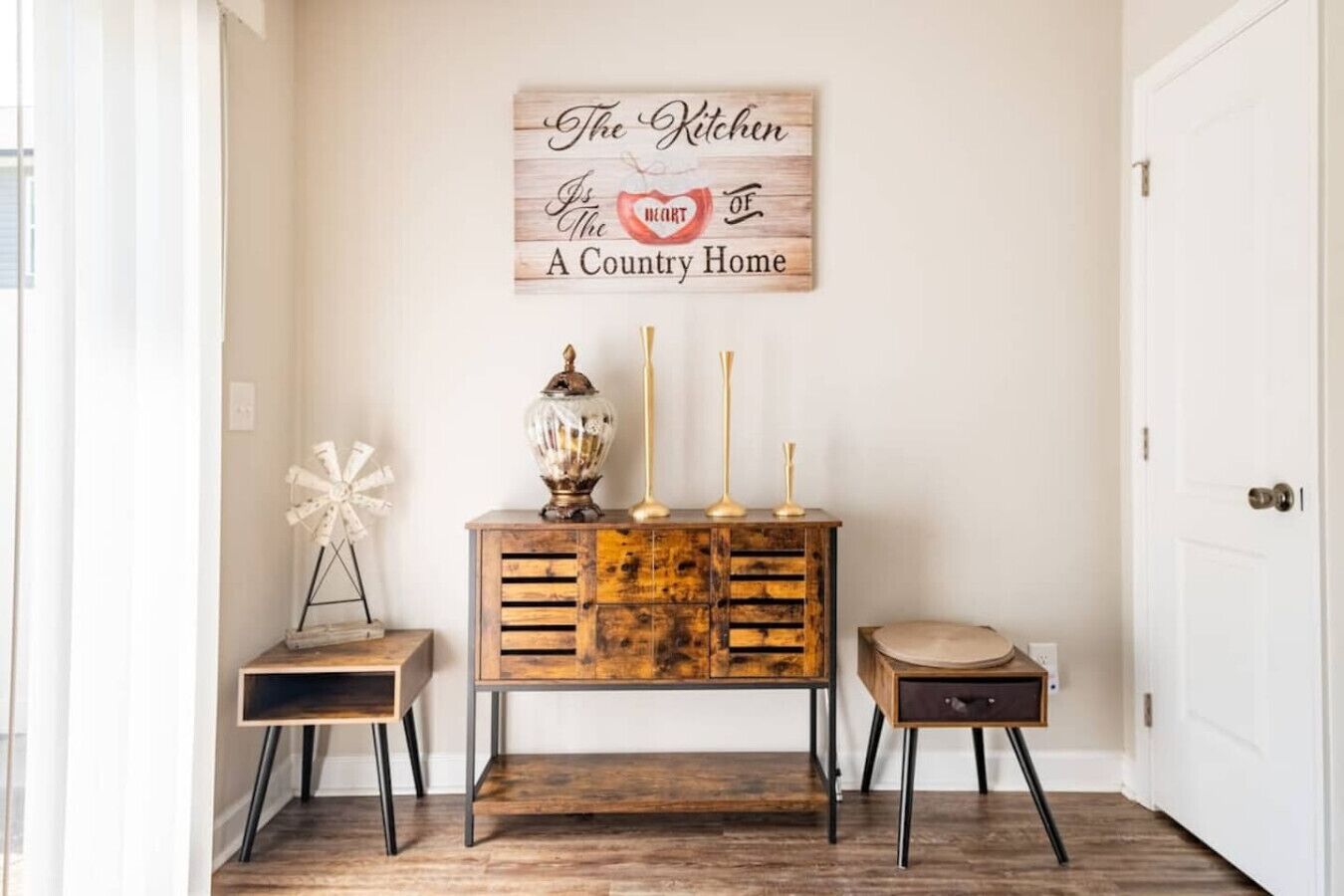 A hallway with a rustic wooden console table, a white windmill decoration, and a sign above stating "The Kitchen is the heart of a country home."