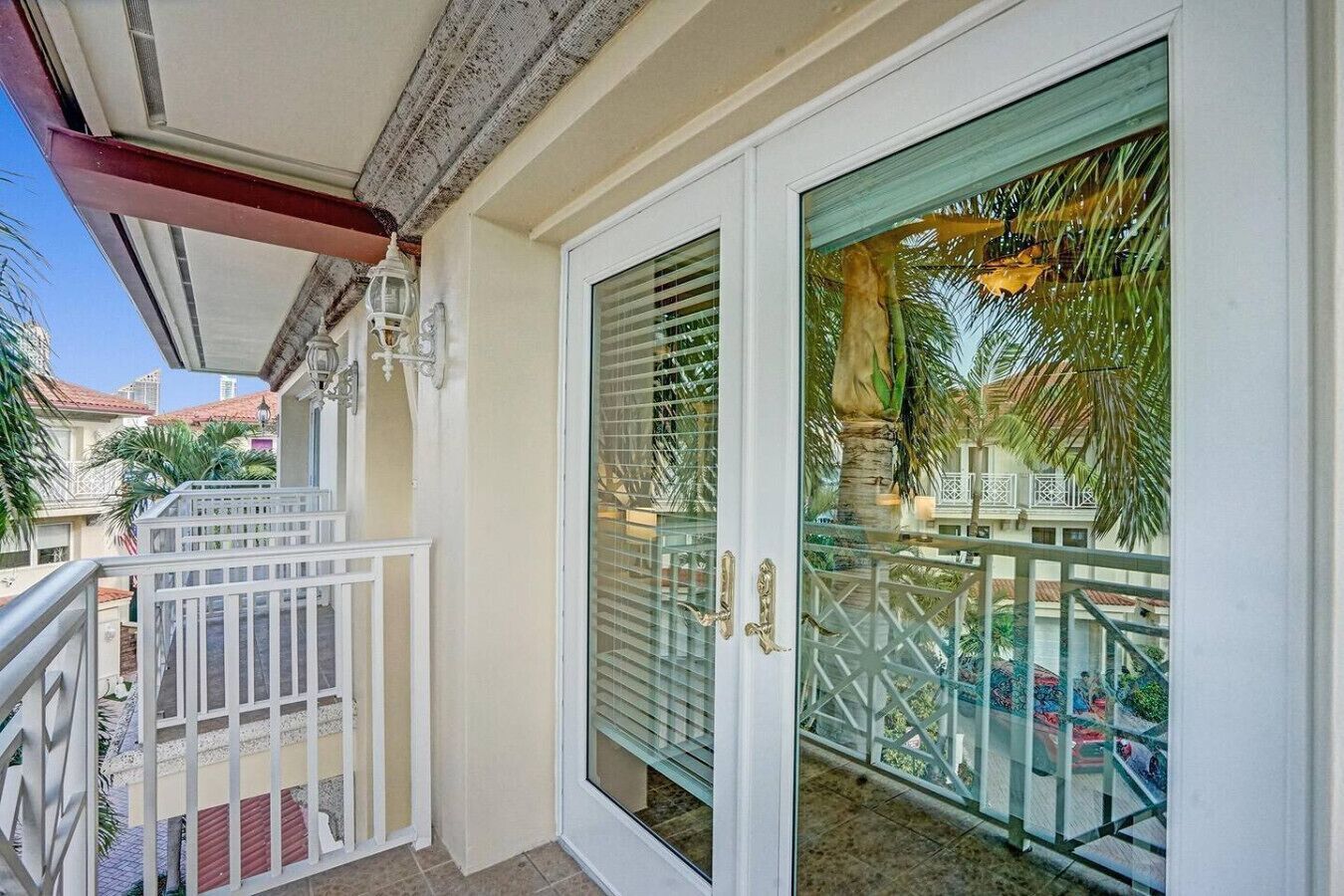Un balcón con barandal blanco, puerta de vidrio con manija metálica y vista a una zona residencial con palmeras y casas.