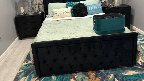 Un dormitorio con una cama tapizada de negro, un suelo con estampado y una mesita de noche con lámpara y objetos decorativos.