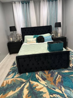 A bedroom with a tufted headboard bed, a patterned rug, and a nightstand with a lamp and decorative items.