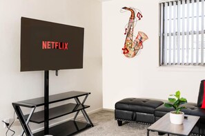 Een woonkamer met een op een standaard gemonteerde televisie, een saxofoon muurschildering, een zwarte leren bank en een kleine tafel met een plant.