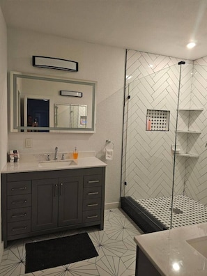 A modern bathroom with a grey vanity, a large mirror, a shower with a herringbone tile pattern, and a black rug on the floor. - 632 4 Large Bedrooms Spacious Private Yard (Dallas)