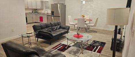 A modern living room with a black leather sofa, a glass coffee table, and a red accent table. There is a kitchen in the background with white cabinets and a stainless steel refrigerator.