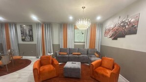 A modern living room with a grey sectional sofa, orange armchairs, and a central ottoman. There is a chandelier, a dining area with a glass table, and a painting on the wall. - 625 3 Large Bedrooms Luxe Artistic Art Vibe (Newark)