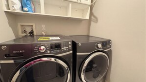 Un área de lavandería con dos lavadoras de carga frontal, un estante con productos de limpieza y una pared blanca con un enchufe.