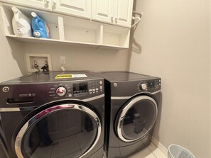 A laundry area with two front-loading washing machines, a shelf with cleaning supplies, and a white wall with a power outlet. - Escape Heaven! Tranquility Guaranteed, Fully Fenced Pet Friendly (Palm Coast)