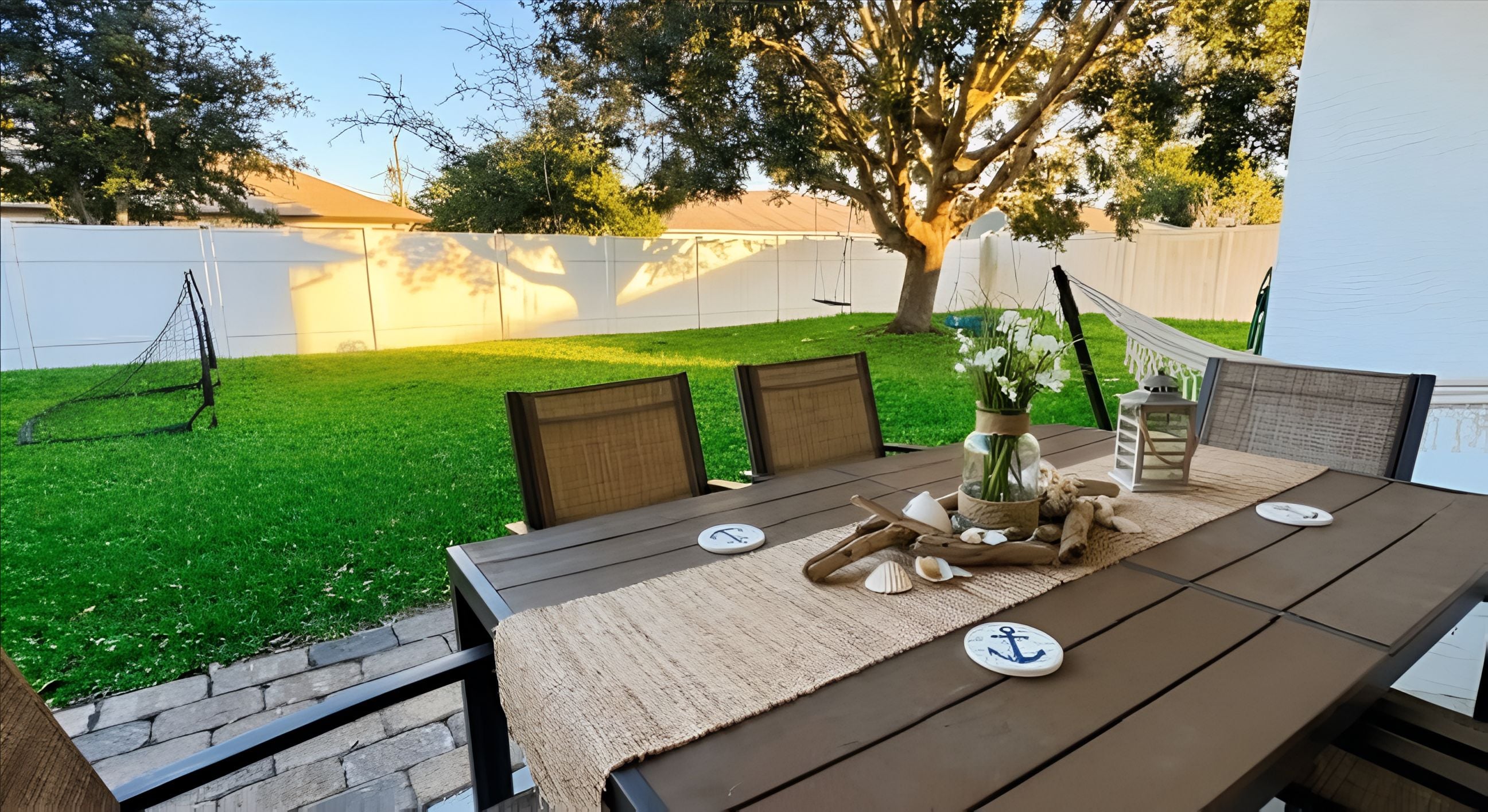 Eine Terrasse mit Tisch für zwei Personen, umgeben von Stühlen, einem kleinen Garten und einem weißen Zaun.