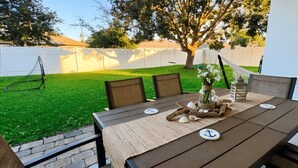 Un patio con mesa lista para dos personas, rodeado de sillas, un jardín pequeño y una cerca blanca.