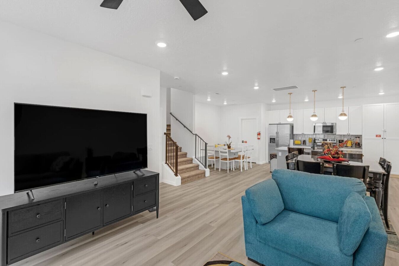 Un soggiorno moderno con un'ampia televisione a schermo piatto, un mobiletto in legno scuro e una poltrona blu. Una scala conduce a una zona pranzo con tavolo e sedie bianche. Si intravede in secondo piano la cucina, con mobili bianchi ed elettrodomestici in acciaio inossidabile.