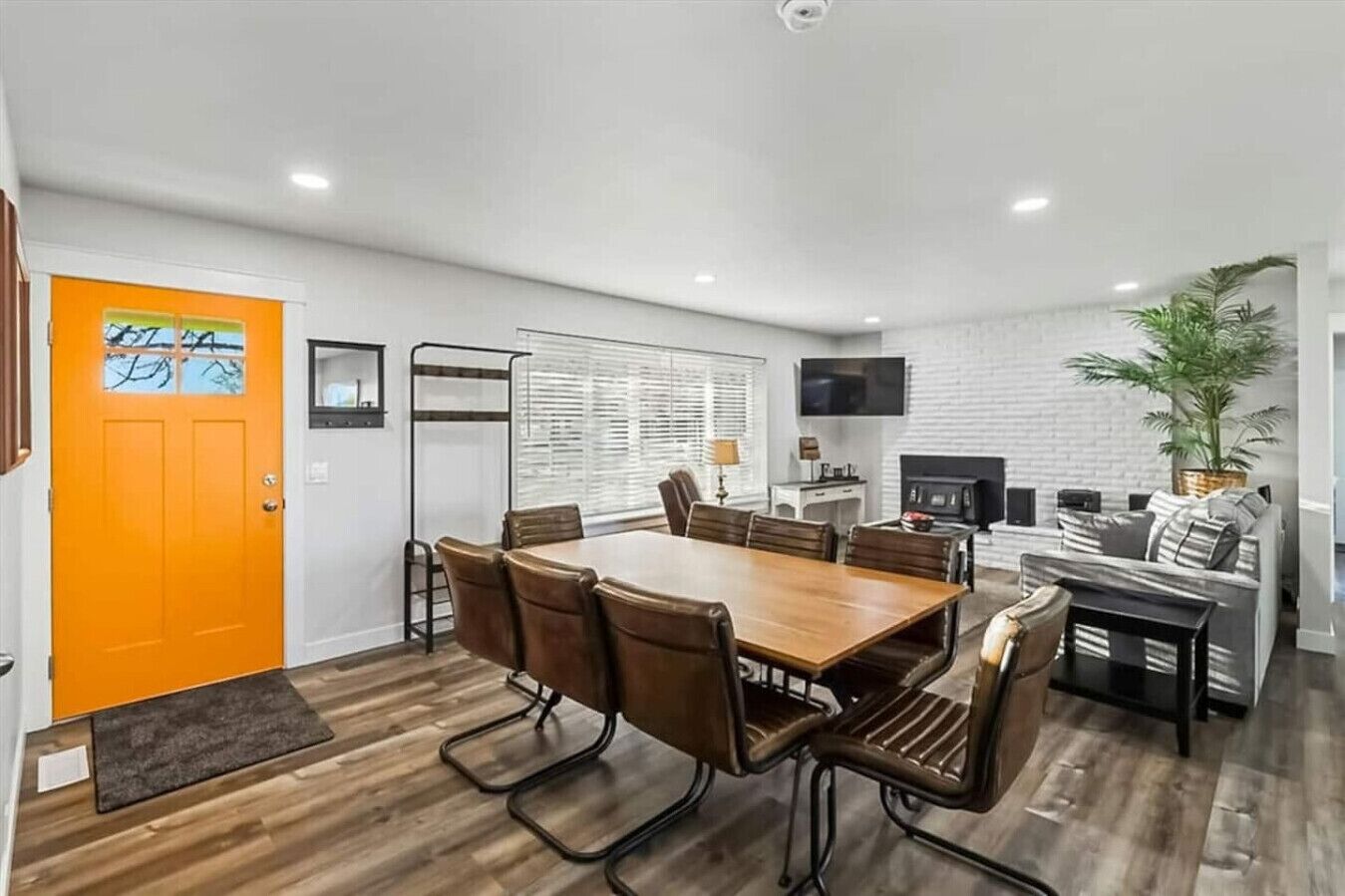 A modern kitchen with a dining area, white brick walls, and a bright orange door.