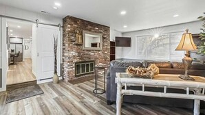 A modern living room with a brick fireplace, a grey sofa, a coffee table with decorative items, and a dining area visible through an open door. - 539 Massive 3 Bedroom Sleeps 10! (Yakima)