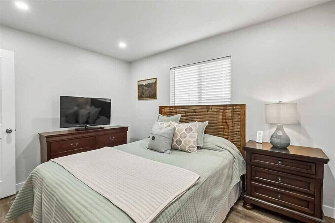 Une chambre à coucher avec un lit, un téléviseur posé sur une commode, une lampe et une fenêtre avec des stores.