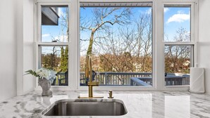 A kitchen with a marble countertop, a stainless steel sink, and a view of a tree and railing through large windows.