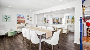 A modern dining area with a long table, white chairs, and a kitchen in the background.