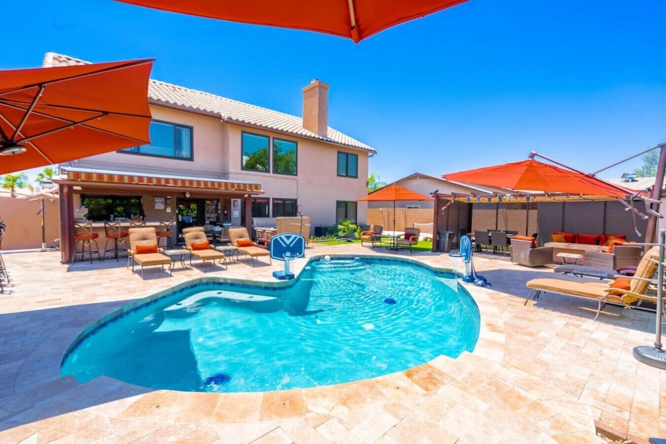 Une piscine entourée de chaises longues et de parasols, avec une terrasse aménagée avec des sièges d’extérieur et un bâtiment en arrière-plan.