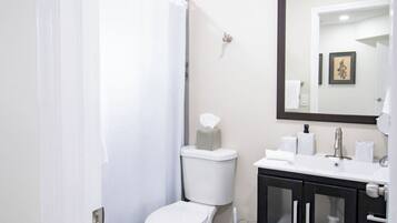 Un baño con inodoro, cortina de ducha, lavamanos con espejo y un cubo de basura.