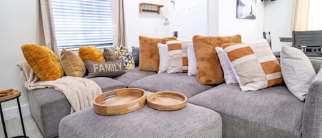A modern living room with a grey sectional sofa, wooden trays, and a 'FAMILY' pillow.