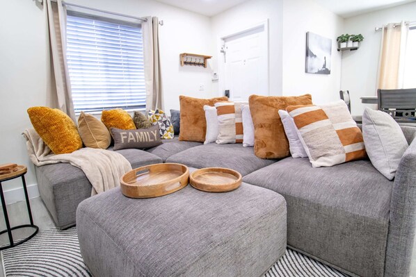 Una sala moderna con un sofá seccional gris, bandejas de madera y un cojín con la palabra 'FAMILY'.
