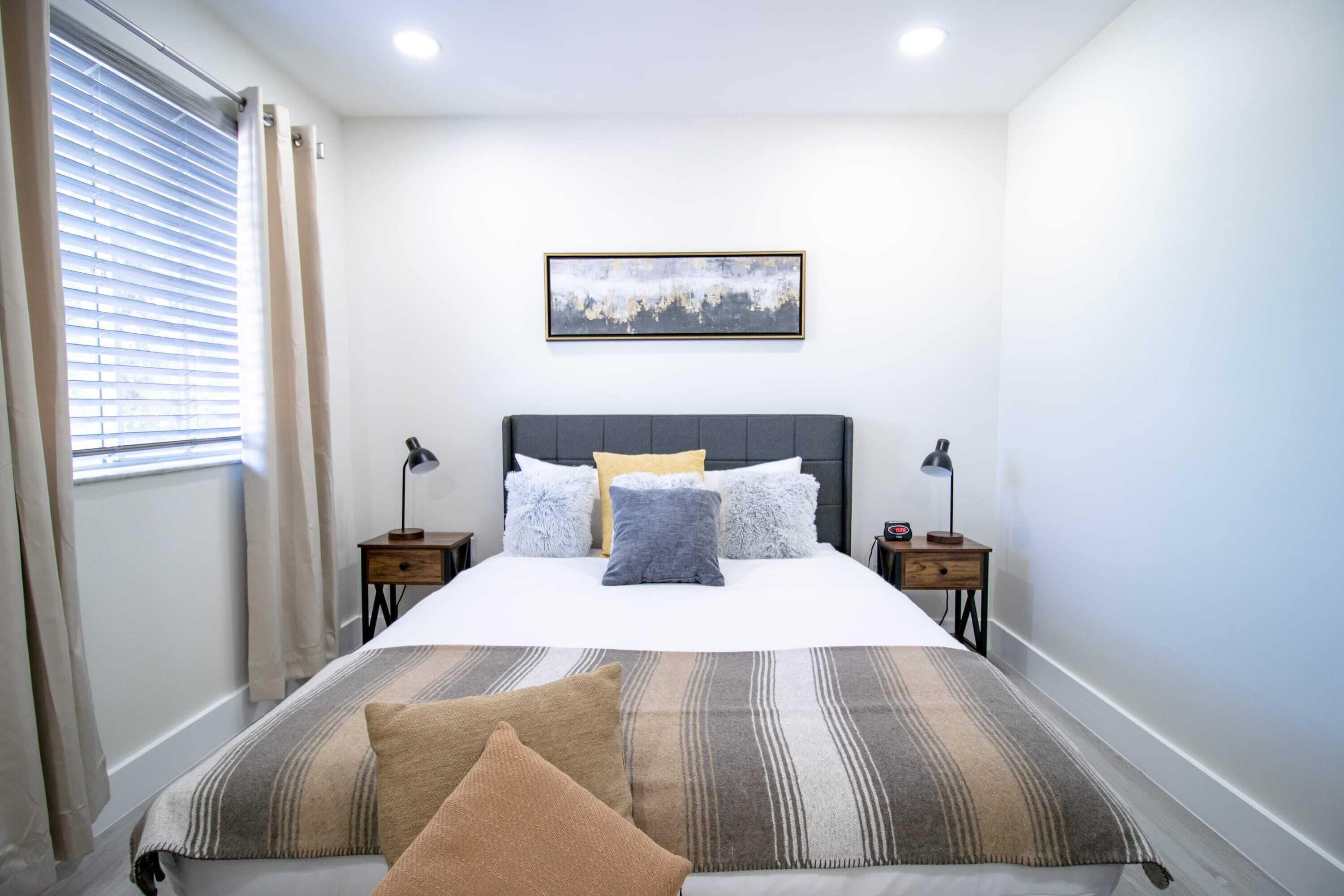 A bedroom with a bed, two bedside tables, a nightstand, a window with curtains, and a wall-mounted picture.