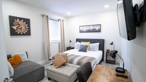 A hotel room with a bed, a desk, a chair, a small ottoman, a nightstand, a lamp, a television, and artwork on the walls. - Good Vibes Oasis - Cozy Townhome With Parking (Cutler Bay)