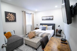 Una habitación de hotel con cama, escritorio, silla, pequeño reposapiés, mesita de noche, lámpara, televisor y obras de arte en las paredes.