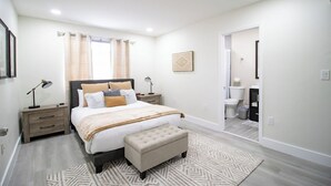 A bedroom with a bed, bedside tables, a nightstand, a bench, a window with curtains, a wall-mounted picture, and a bathroom visible through an open door. - Good Vibes Oasis - Cozy Townhome With Parking (Cutler Bay)