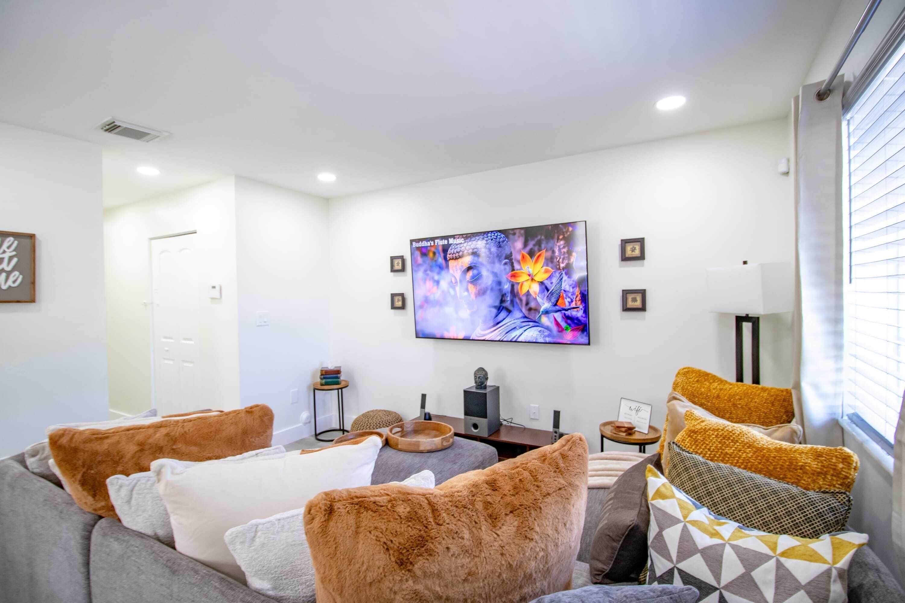 Una sala de estar moderna con un televisor de pantalla plana, un sofá con cojines estampados, una mesita auxiliar pequeña y un elemento decorativo en la pared.