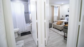 A modern bathroom with a toilet, shower, and a white door leading to a bedroom with a bed and a desk. - Good Vibes Oasis - Cozy Townhome With Parking (Cutler Bay)