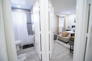 Un baño moderno con inodoro, ducha y una puerta blanca que da a un dormitorio con cama y escritorio.