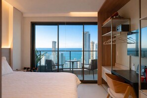 Ein Hotelzimmer mit einem Bett, einem kleinen Tisch, einem Stuhl, einem Schreibtisch mit Computer und einem großen Fenster mit Blick auf die Stadt.