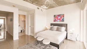 A modern bedroom with a bed, bedside tables, a painting, and a bathroom in the background. - Hang Ten- Large Townhome, lux Amenities, Parking (Miami)
