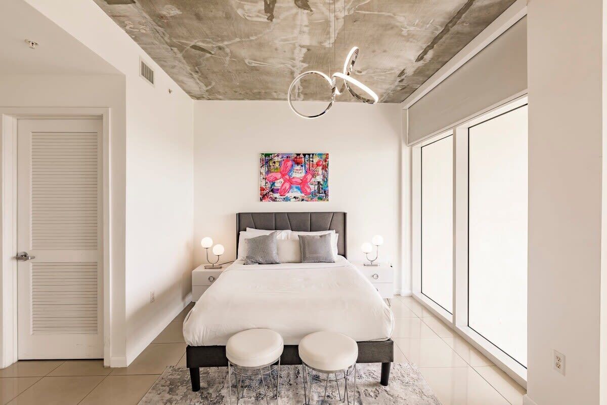 Une chambre avec un lit, deux tables de chevet, une grande fenêtre et un plafond en béton apparent.