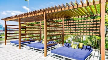 Un salón en la azotea con pérgolas de madera, asientos acolchados azules y un fondo de vegetación.