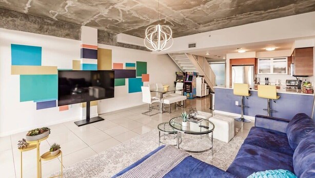 A modern living room with a flat-screen TV, a sofa, a coffee table, and a staircase leading to the kitchen. - Hang Ten- Large Townhome, lux Amenities, Parking (Miami)