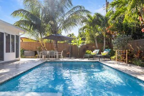 A swimming pool with a patio area, surrounded by palm trees and a wooden fence.