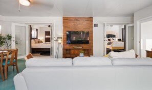 A modern living room with a large flat-screen TV, a wooden accent wall, a sofa, and a dining area with chairs.