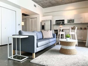 A modern living room with a grey sofa, a small white side table, a wicker coffee table, and a kitchen area with white cabinets and appliances. - Center Stage- Downtown, Parking, Pool, Hot Tub (Miami)