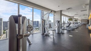 A modern gym with various exercise equipment, large windows offering a city view, and a clean, spacious layout.